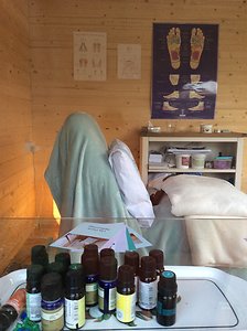 The cabin from the other end: a client resting under a pale green blanket, a shelf of essential-oil bottles in the foreground and reflexology charts pinned to the pine wall.