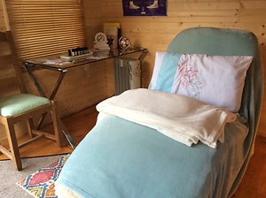 Inside Lila's garden cabin: a therapy couch made up in sage green with a cream towel and an embroidered cushion, pine-panelled walls, a bamboo blind and a small glass side table with candles.